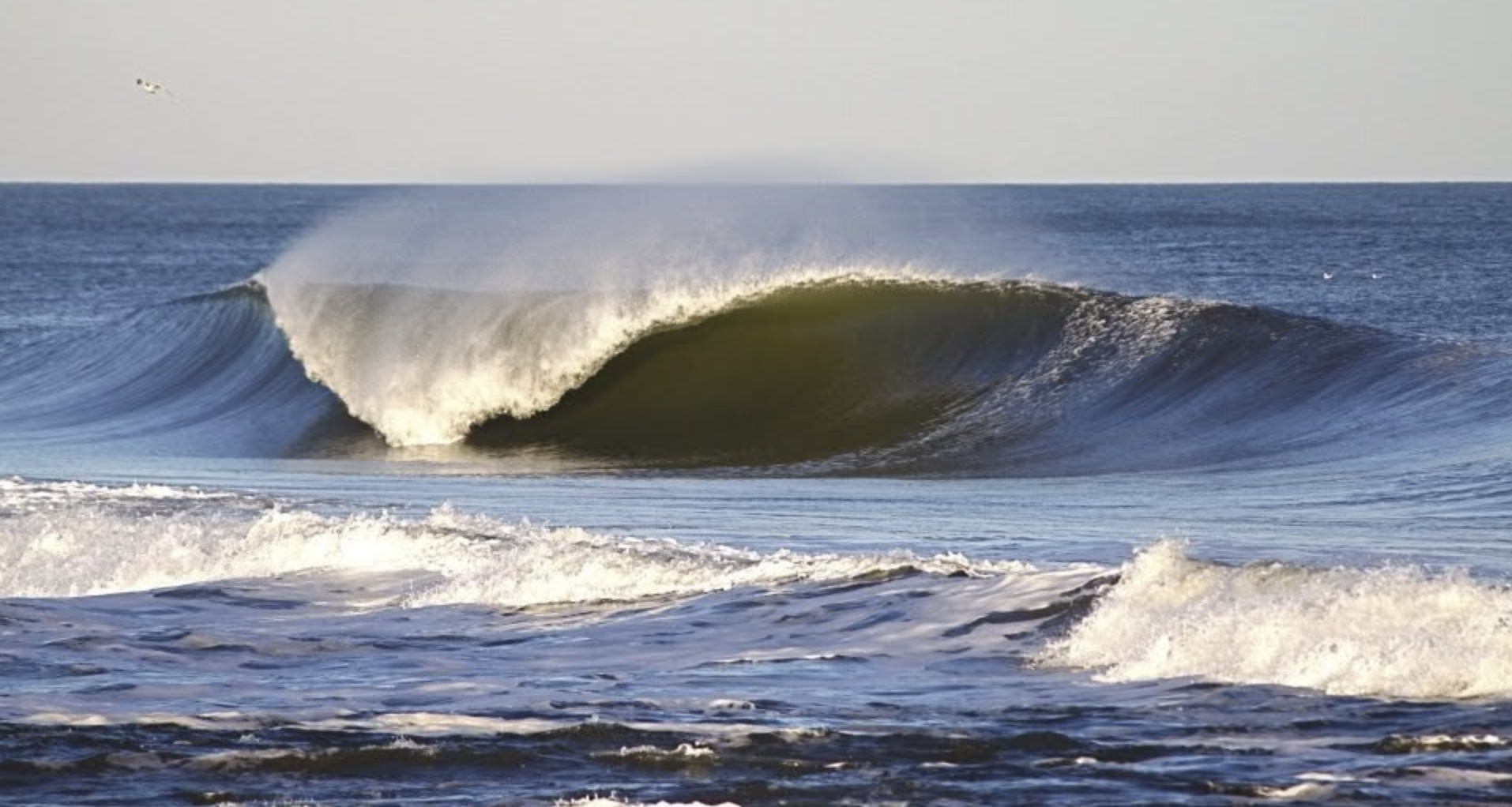 An ocean wave breaking into a hollow barrel, with spray blowing off the crest.